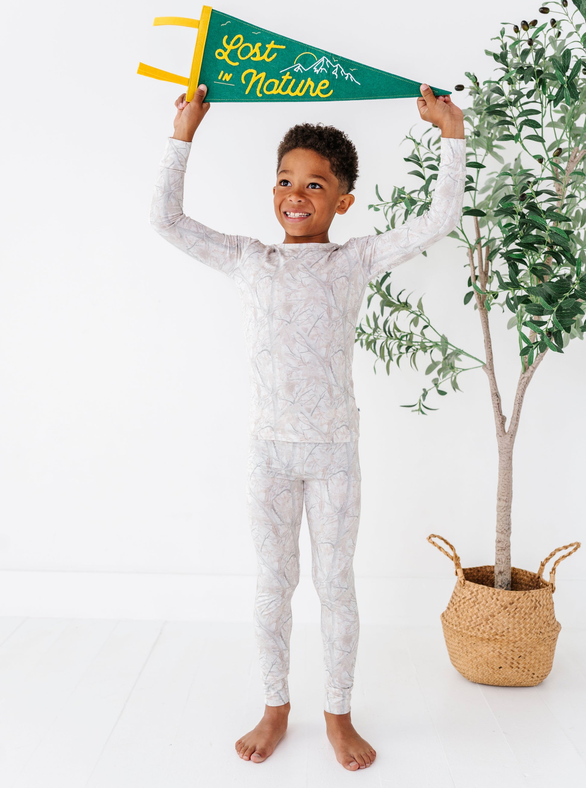 Child in tree camo patterned pajamas holding a 'Lost in Nature' flag with a plant in the background.