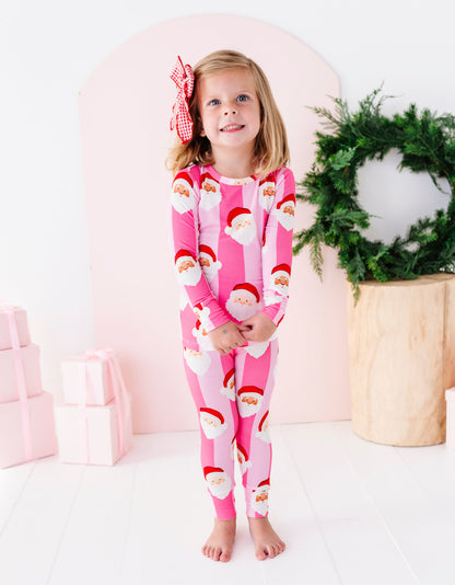 A young girl stands barefoot in a studio setting wearing a pink long-sleeve bamboo pajama set with a repeating Santa Claus face print. She has a red and white bow in her hair, with a pink arch backdrop, wrapped gifts, and a green wreath in the background.