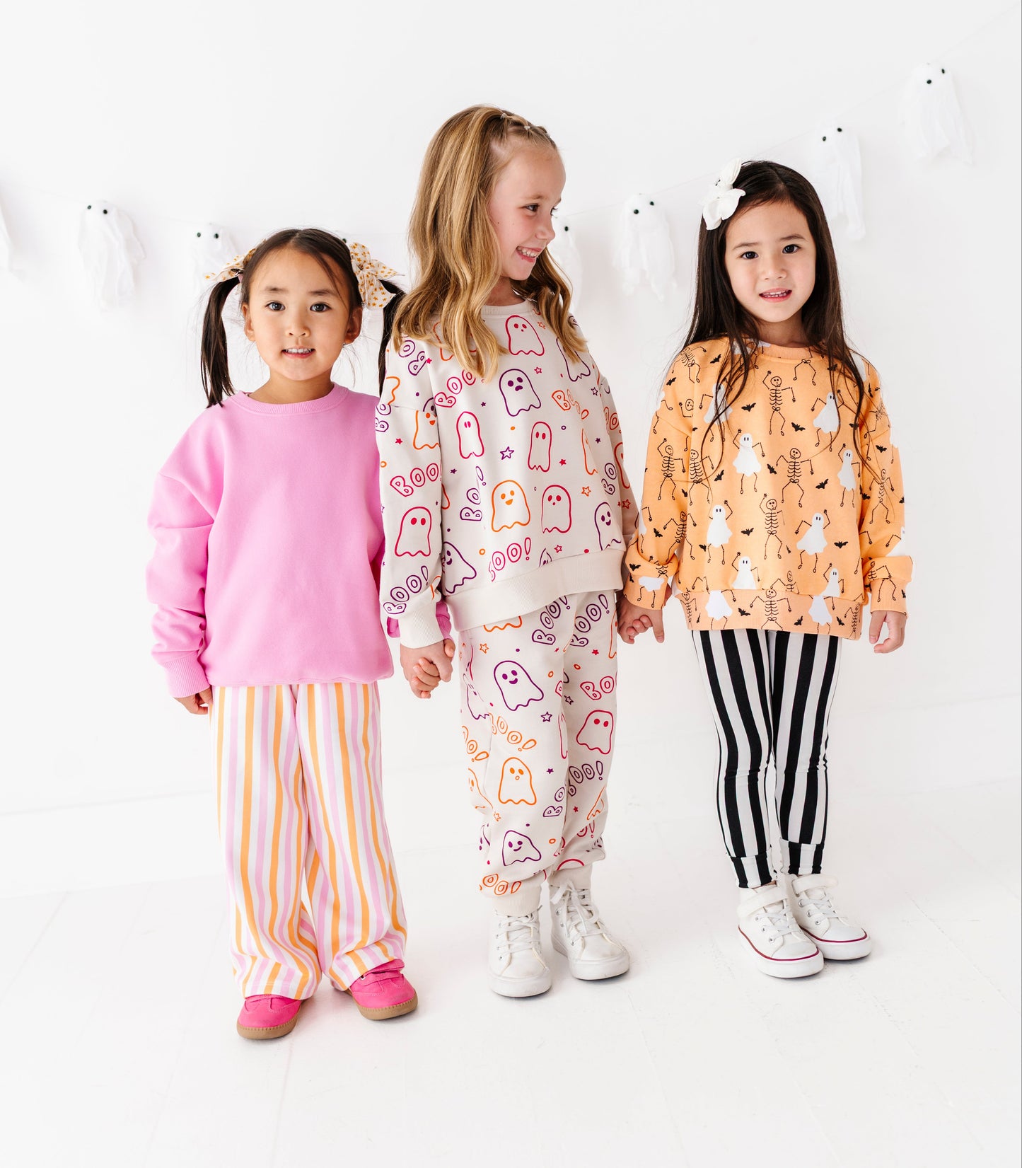 Three young girls modeling matching pajama sets in a bright white studio. The girl on the left wears a pink long-sleeve top with orange and white striped pants.