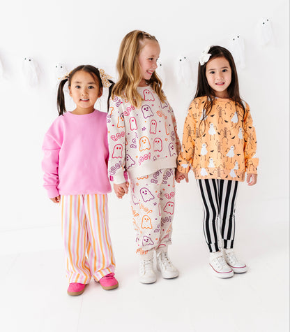 Three young girls modeling matching pajama sets in a bright white studio. The girl on the left wears a pink long-sleeve top with orange and white striped pants.