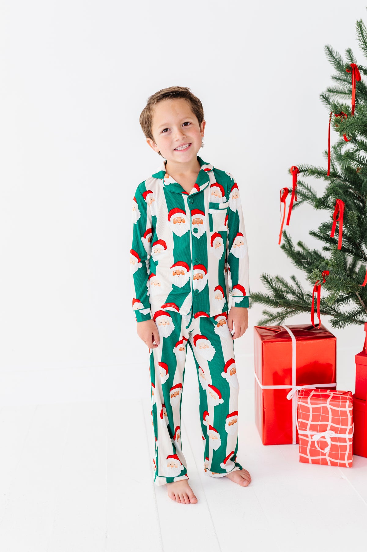 A young boy stands barefoot in a white studio setting wearing green bamboo pajamas with a Santa Claus face pattern. To his right is a decorated Christmas tree with red ribbons and wrapped presents.