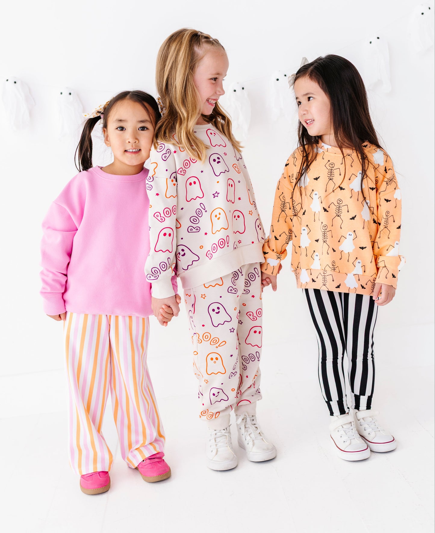 Three young girls wearing matching halloween-themed sets stand together against a white studio background decorated with white ghosts. The girl on the left wears a pink sweatshirt and yellow-and-white striped pants.