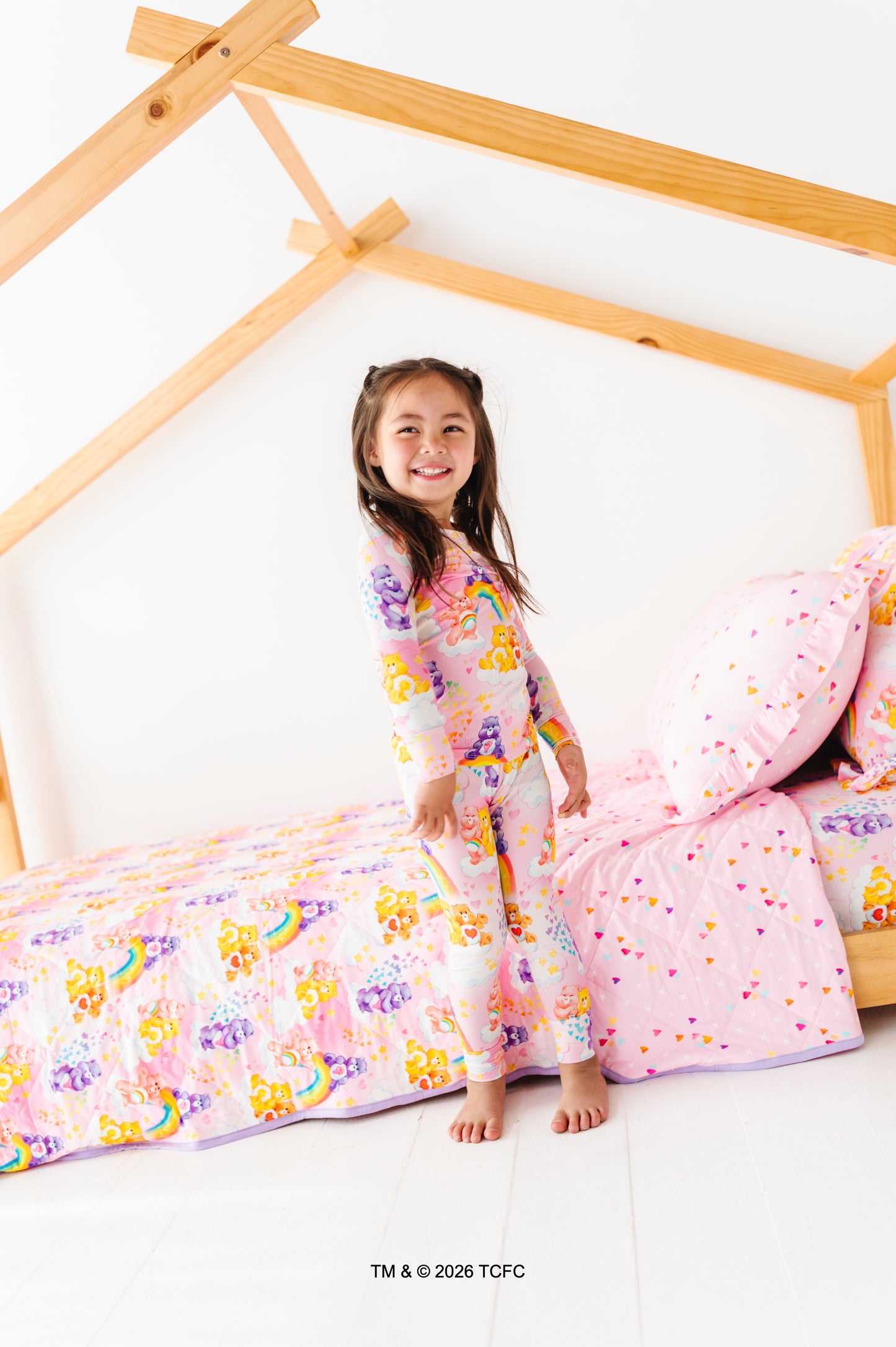 Young girl in a colorful bamboo Care Bears™ pajama set sitting on a bed with a wooden headboard.