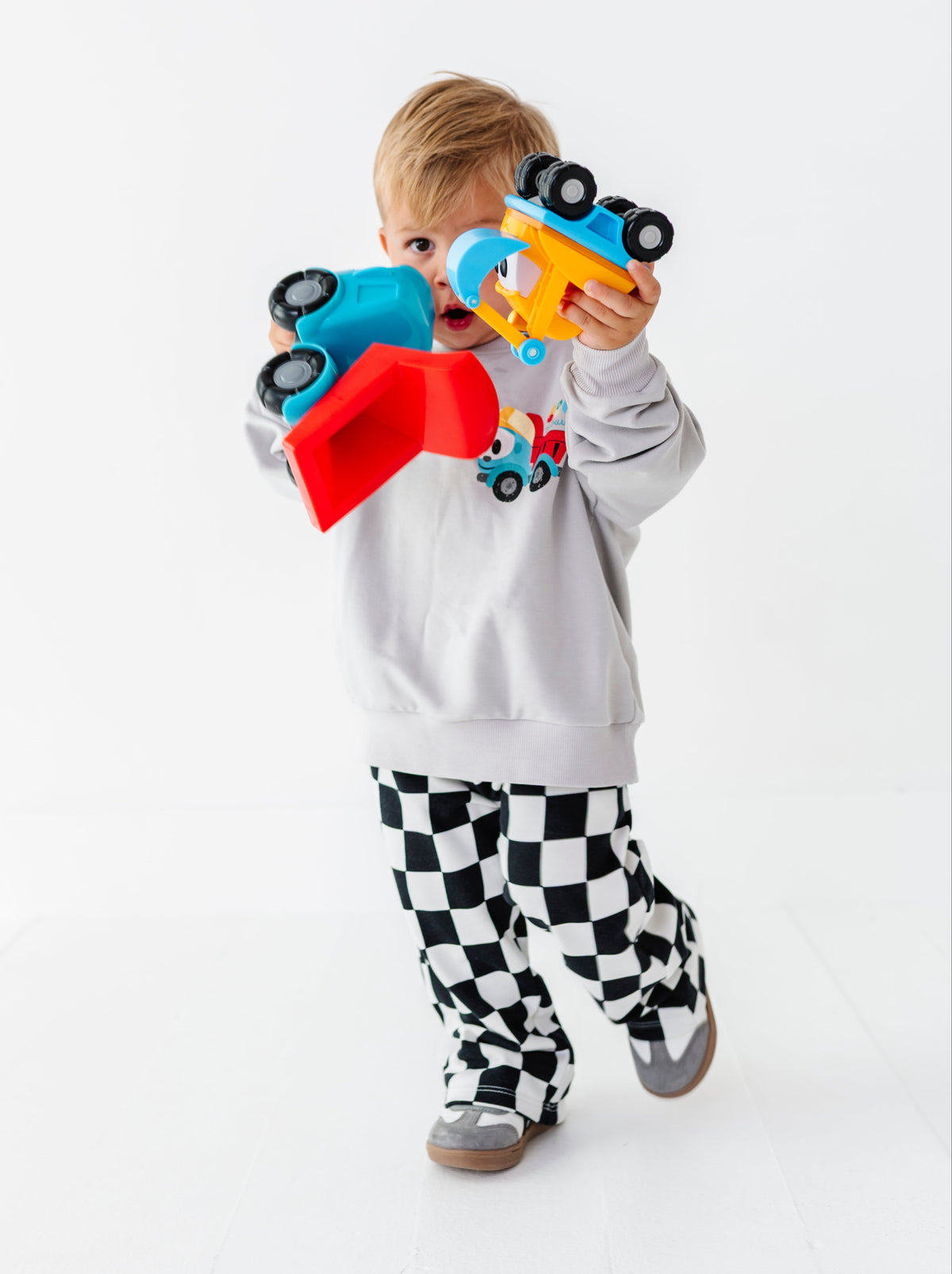 A toddler stands against a white studio background holding two toy dump trucks, one blue with a red scoop and one yellow with blue details. The child wears a light gray sweatshirt with a truck graphic and black-and-white checkered pants.