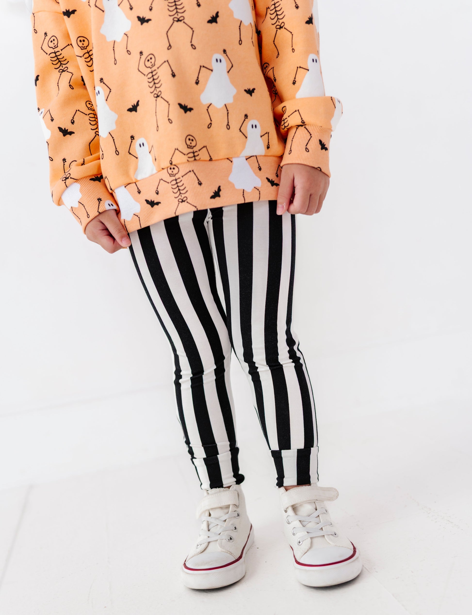 A child wearing a peach-orange long-sleeve sweatshirt with a white and black skeleton and ghost pattern, paired with black and white vertically striped leggings and white low-top sneakers.