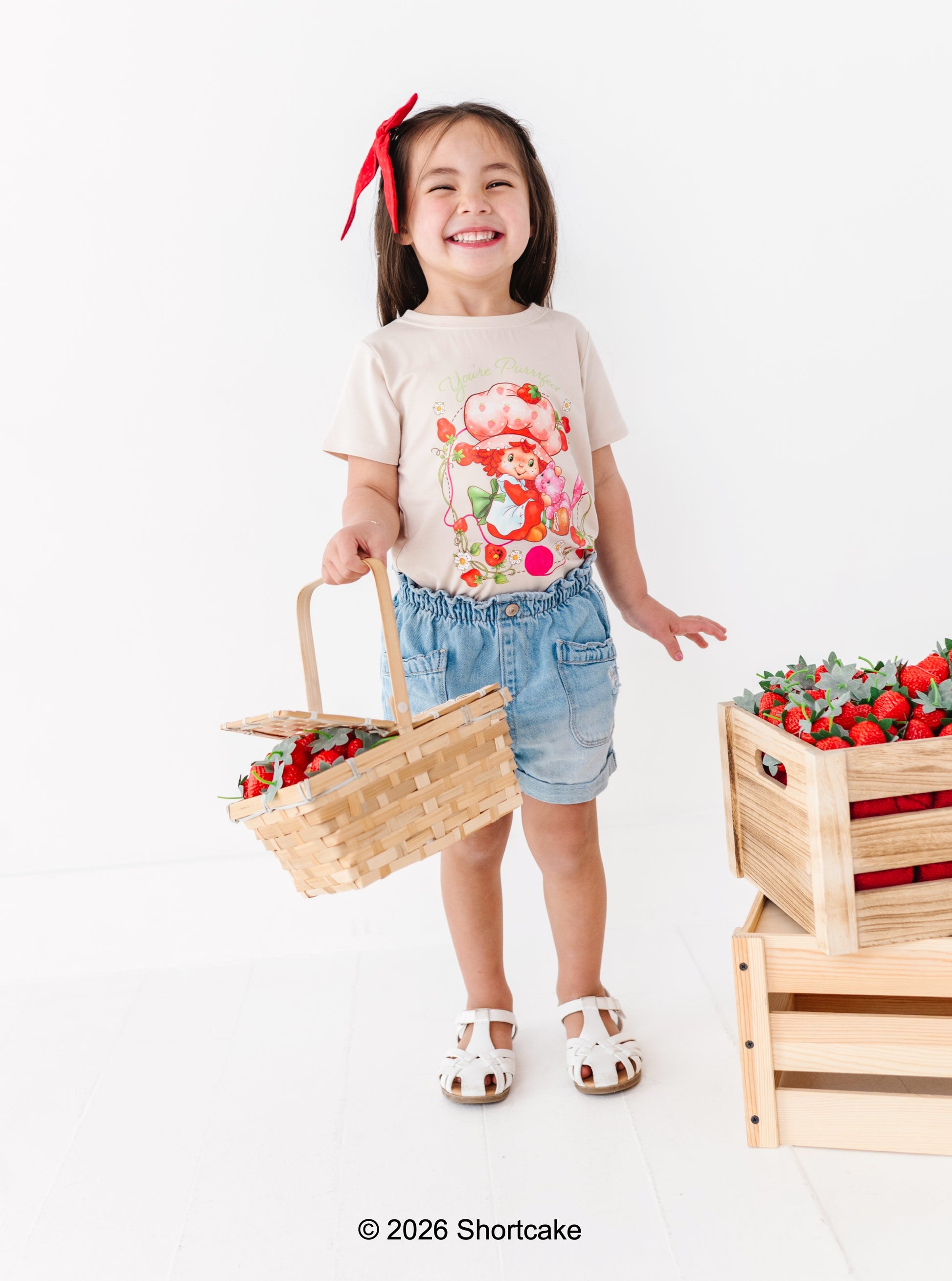 Child holding a basket of strawberries with a crate of strawberries on a white background and wearing a Strawberry Shortcake™ tee 