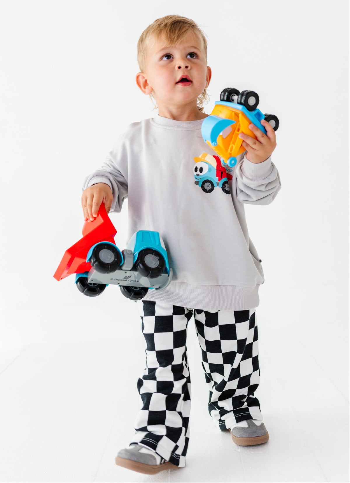 A toddler boy wearing a light gray sweatshirt and black-and-white checkered pajama pants, holding three colorful toy cars against a plain white studio background.