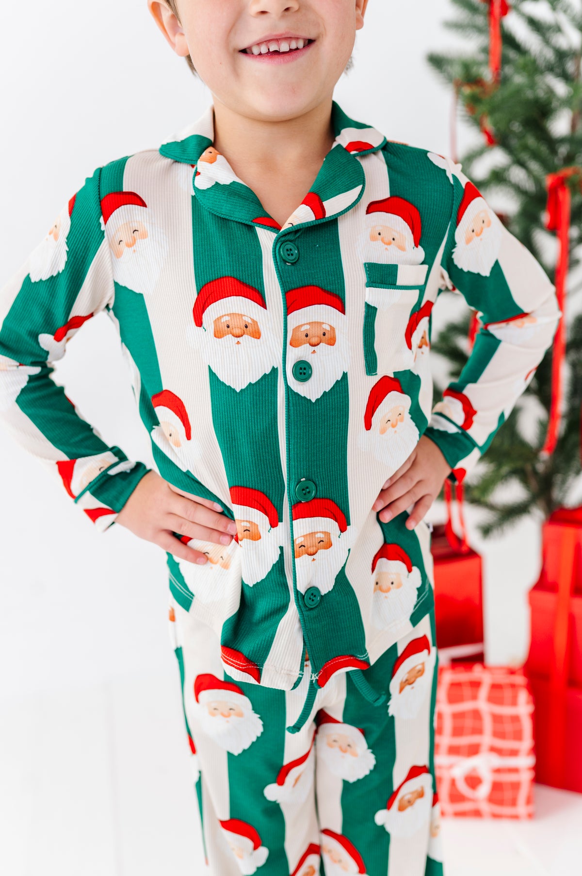 A child wearing a green and white long-sleeve bamboo pajama set with an all-over Santa Claus face print, standing with hands on hips in front of a Christmas tree and wrapped gifts.
