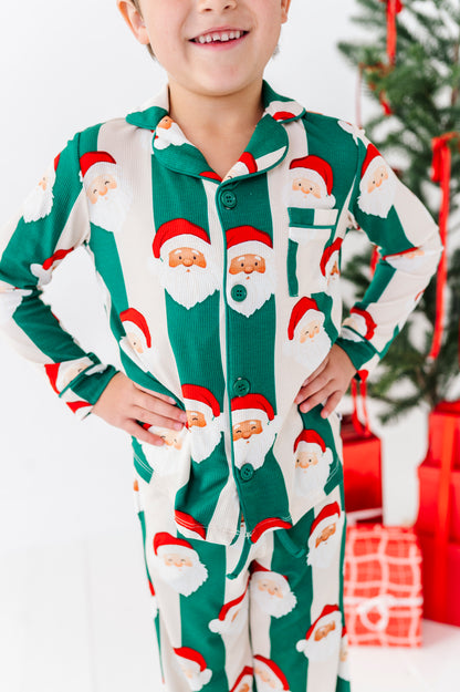 A child wearing a green and white long-sleeve bamboo pajama set with an all-over Santa Claus face print, standing with hands on hips in front of a Christmas tree and wrapped gifts.
