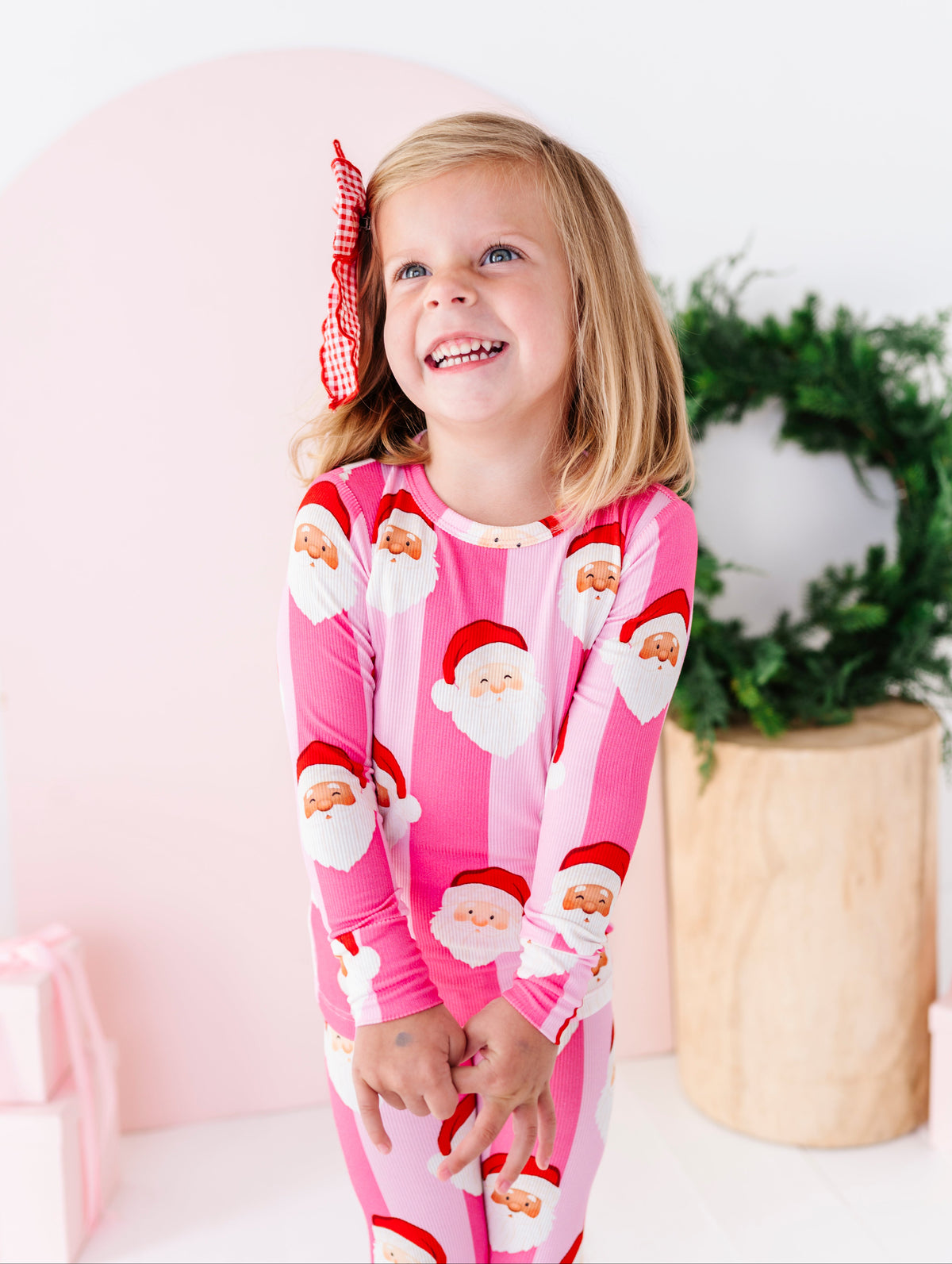 A young girl with blonde hair wearing pink and white striped bamboo pajamas with a Santa Claus pattern, standing in a brightly lit room with a pink arch, a green wreath, and a wooden planter.