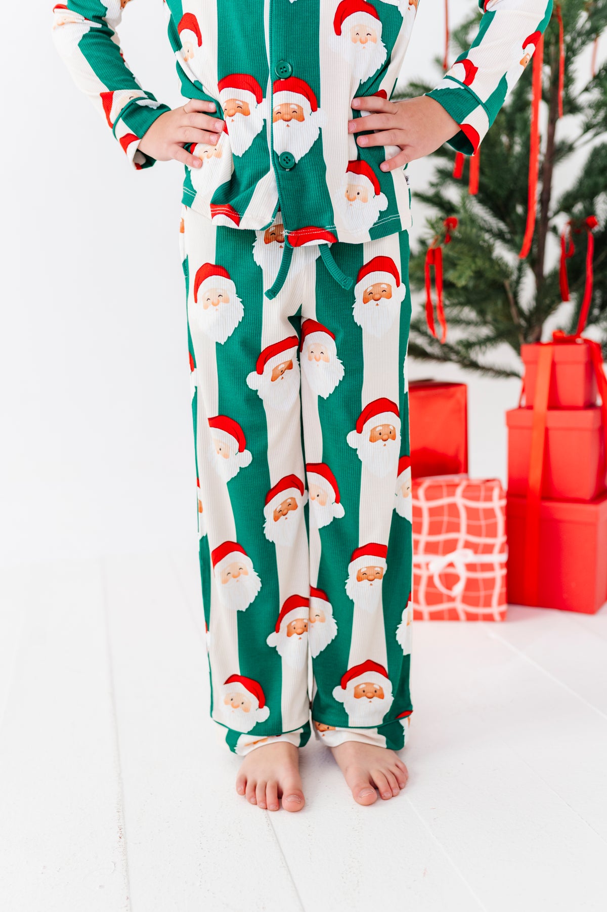 A person wearing festive Christmas bamboo pajamas with a green and white striped Santa Claus print, standing barefoot in a brightly lit room with wrapped gifts and a Christmas tree in the background.