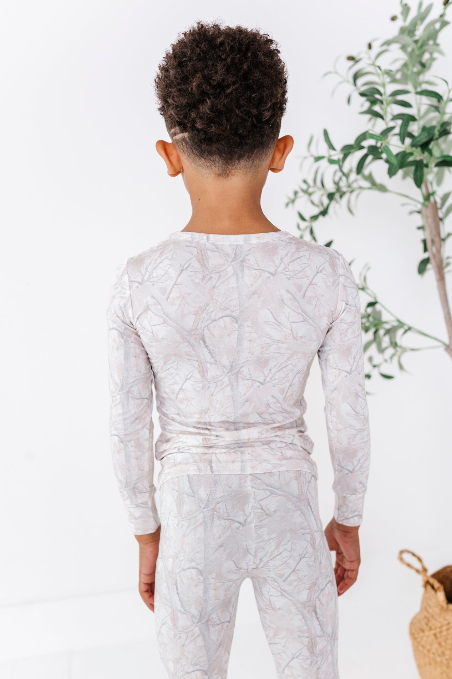 Child wearing a tree camo patterned outfit standing against a light background with a plant in the corner.