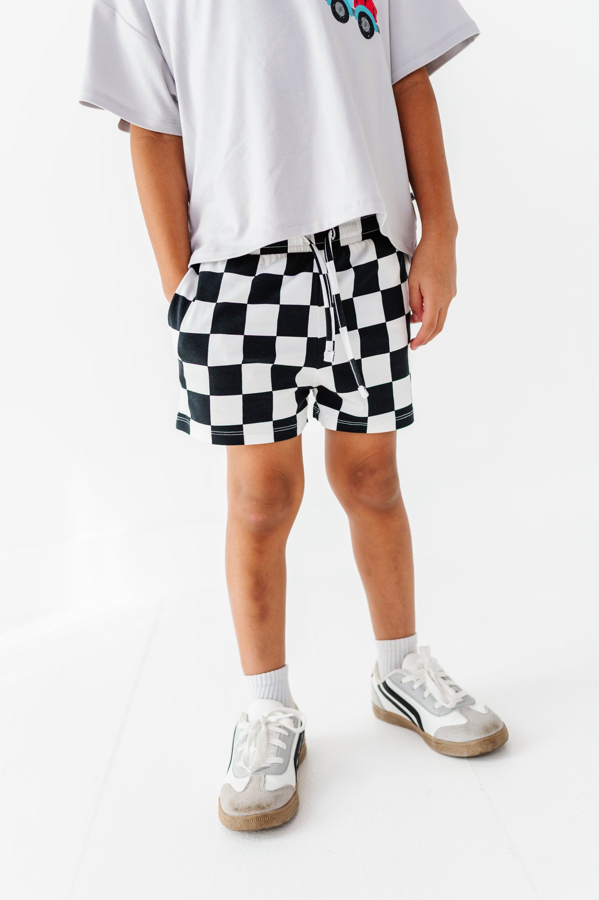 A child wearing black and white checkered shorts, a white t-shirt, and white sneakers, standing against a plain white background.