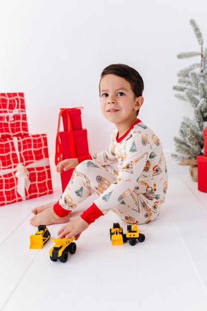 A young boy wearing festive Christmas-themed bamboo pajamas sits on a white floor next to a small decorated tree and wrapped presents, playing with toy construction vehicles.