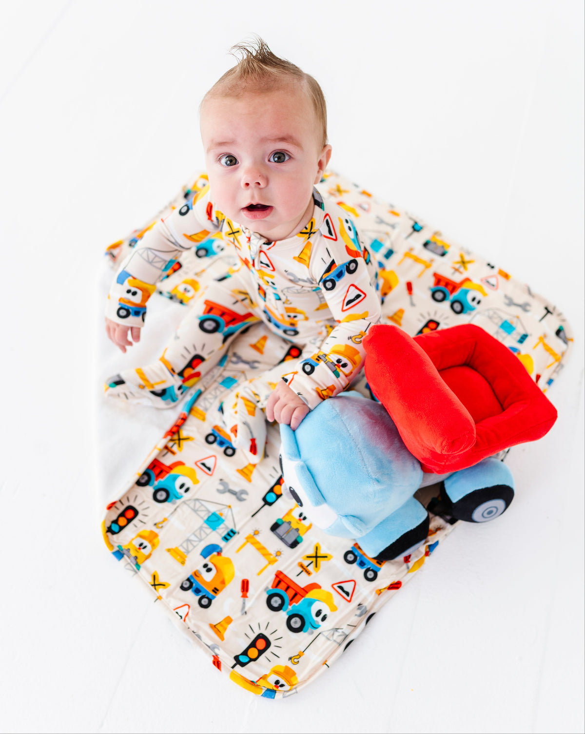 A baby wearing a matching construction-themed onesie lies on a white blanket with a red car seat cushion and a blue plush toy on a white studio background.