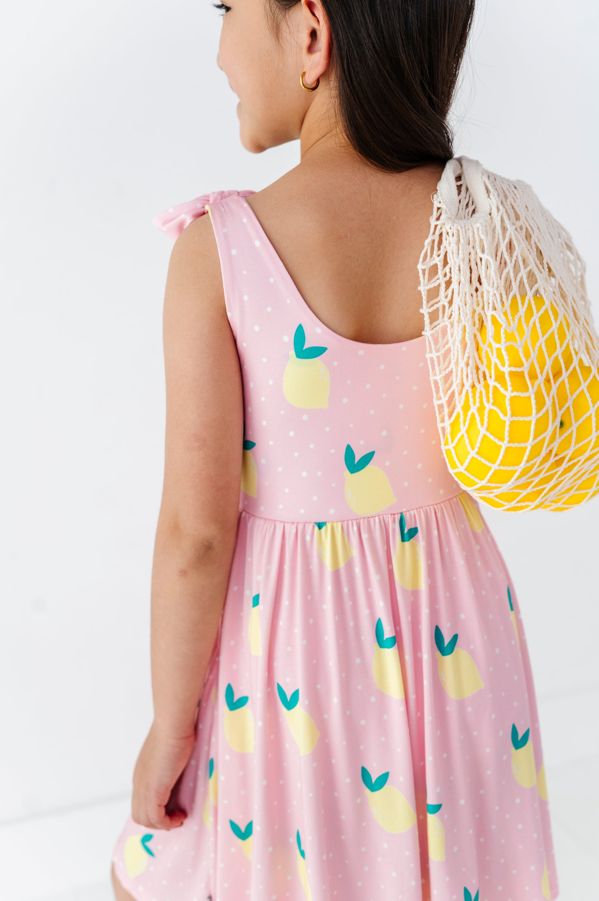 Child wearing a pink dress with lemon patterns, holding a yellow mesh bag.