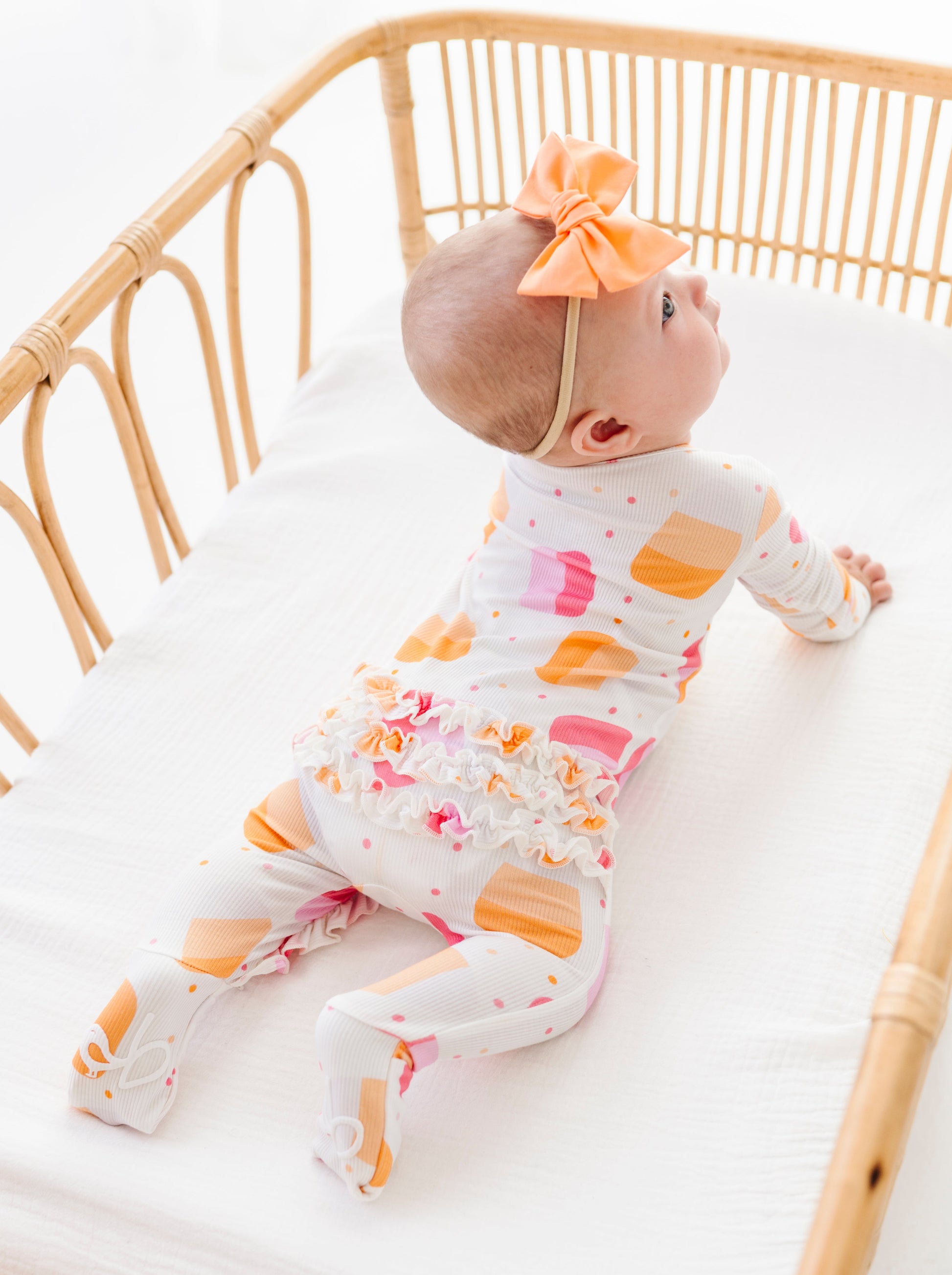 A baby lying on their stomach in a rattan bassinet, wearing a white footed one-piece with orange and pink popsicle prints, a white ruffled trim, and an orange bow headband.