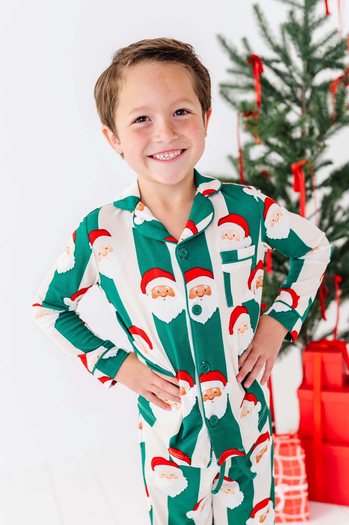 A smiling young boy wearing a green and white Santa Claus patterned long-sleeve bamboo pajama set, standing in front of a decorated Christmas tree and wrapped gifts.