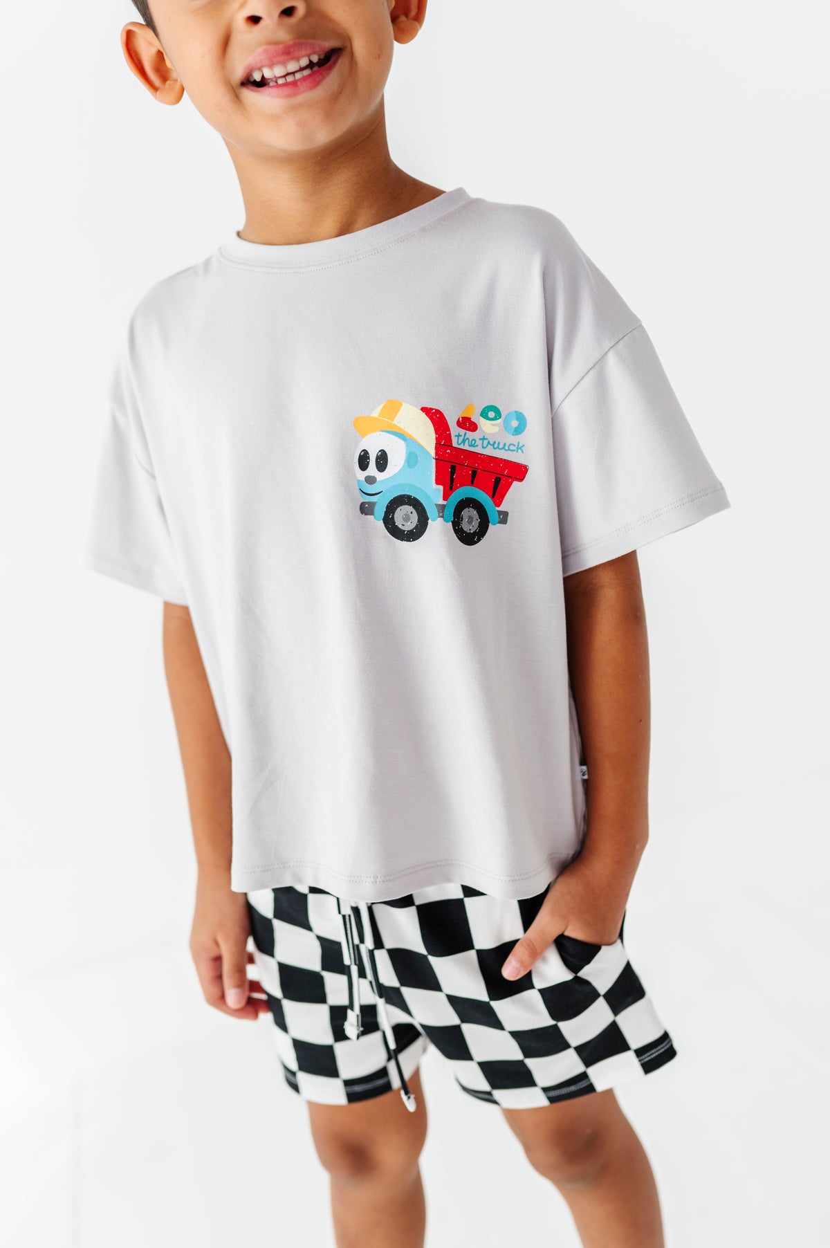 A smiling young boy wearing a white short-sleeve t-shirt with a colorful cartoon dump truck graphic on the chest, paired with black and white checkered shorts, against a plain white background.