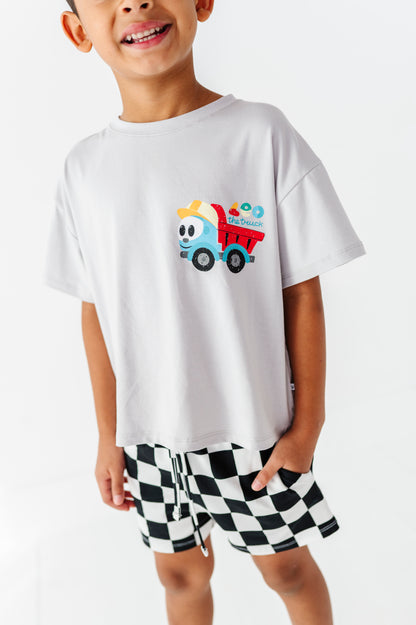 A smiling young boy wearing a white short-sleeve t-shirt with a colorful cartoon dump truck graphic on the chest, paired with black and white checkered shorts, against a plain white background.