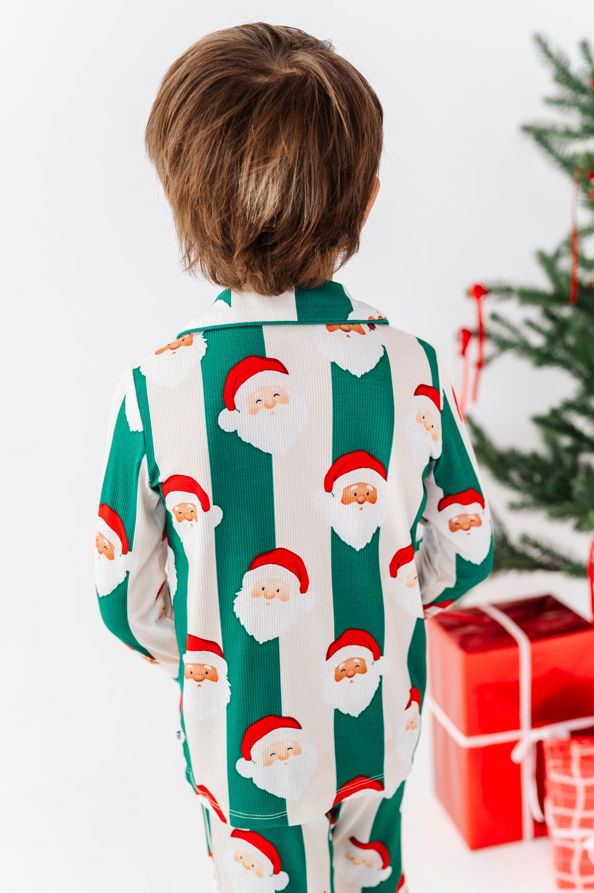 A child viewed from behind wearing green and white striped bamboo pajamas with a Santa Claus face print, standing in front of a Christmas tree and wrapped presents.