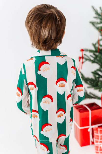 A child viewed from behind wearing green and white striped bamboo pajamas with a Santa Claus face print, standing in front of a Christmas tree and wrapped presents.