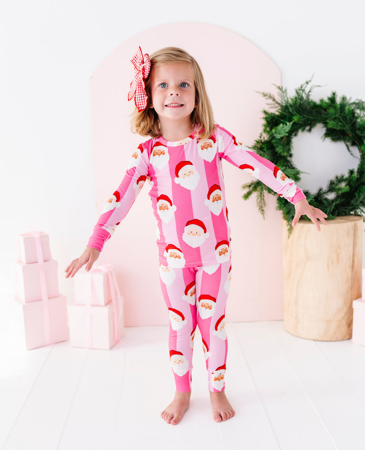 A young child wearing a pink long-sleeve bamboo pajama set with an all-over Santa Claus face print, standing in a brightly lit indoor setting.