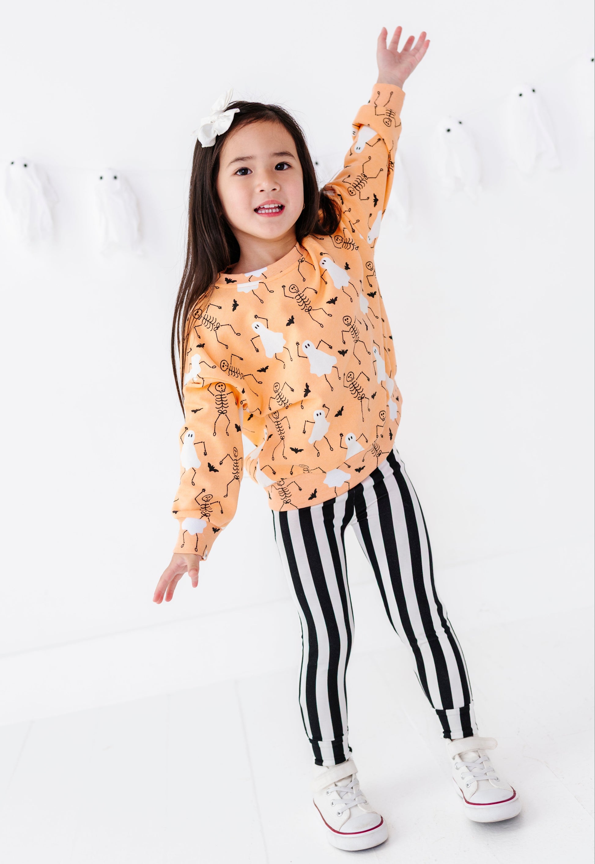 A young girl posing in a bright white studio, wearing a long-sleeve orange sweatshirt with a black and white animal print, black and white vertically striped leggings, and white sneakers.