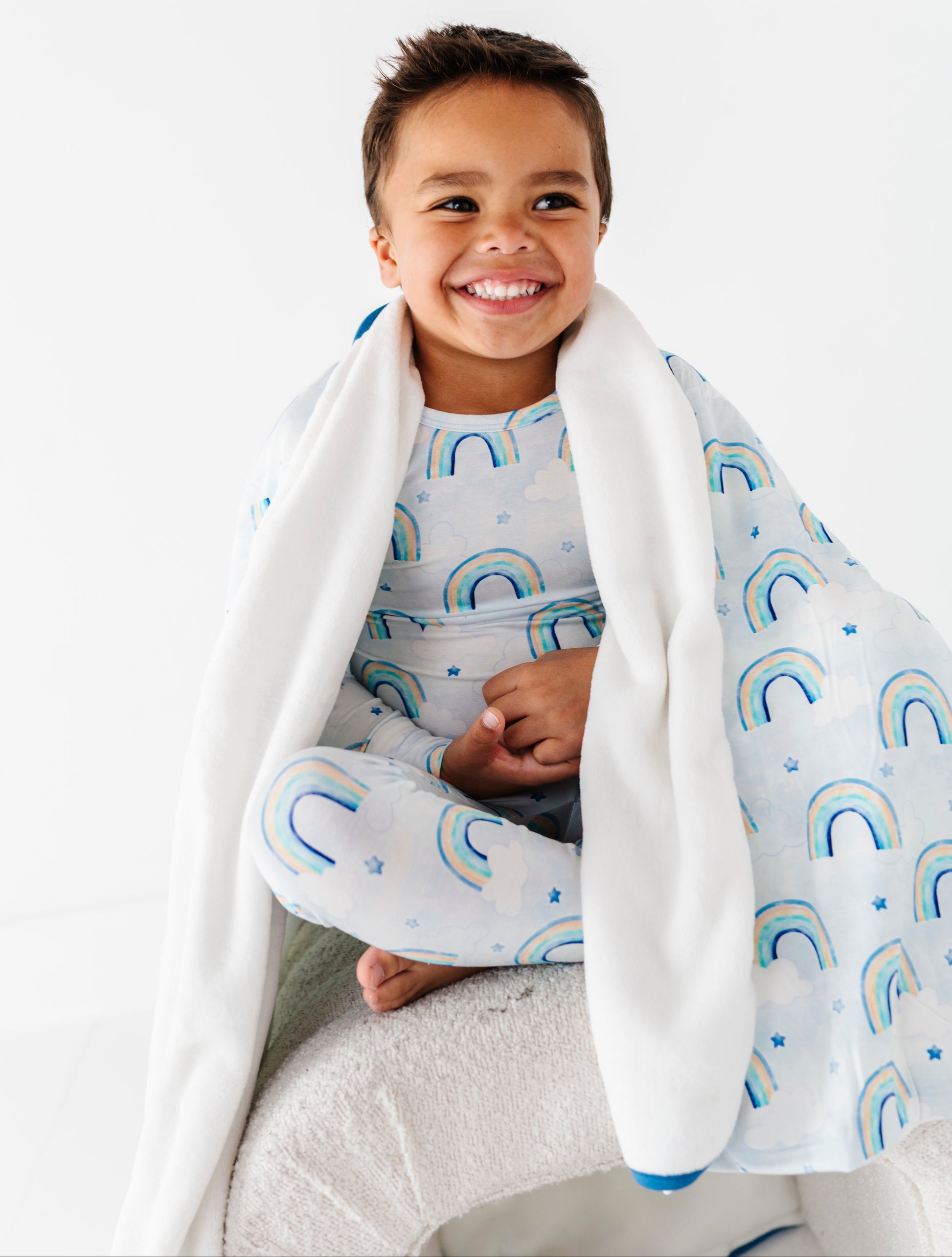 Child wrapped in a bamboo plush blanket with pastel watercolor rainbow patterns, sitting on a white surface.