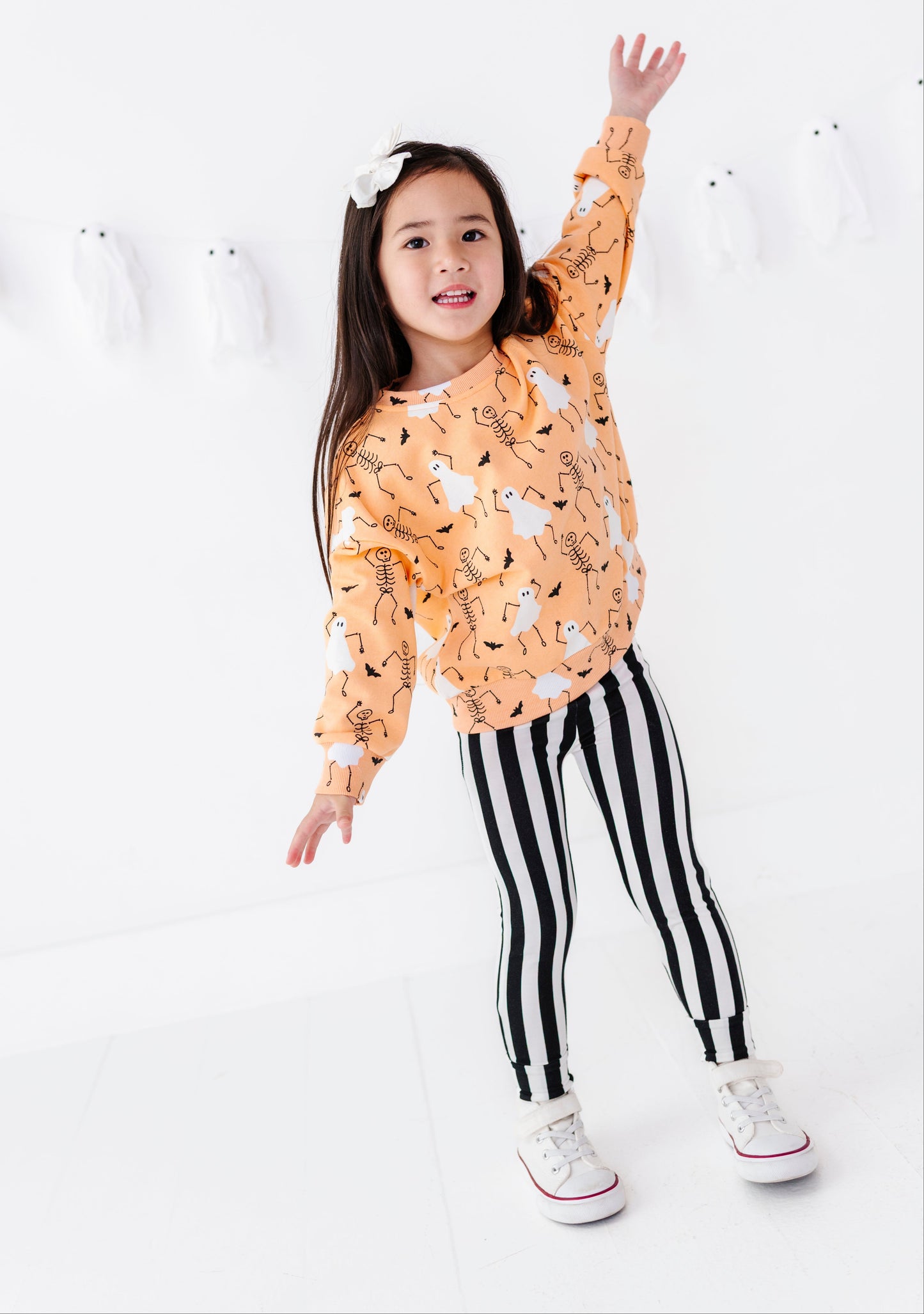 A young girl posing in a bright white studio, wearing a long-sleeve orange sweatshirt with a black and white animal print, black and white vertically striped leggings, and white sneakers.