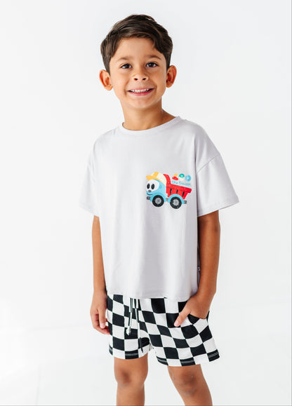 A young boy smiling, wearing a white crewneck t-shirt with a colorful cartoon dump truck graphic on the chest, paired with black and white checkered shorts, against a plain white background.