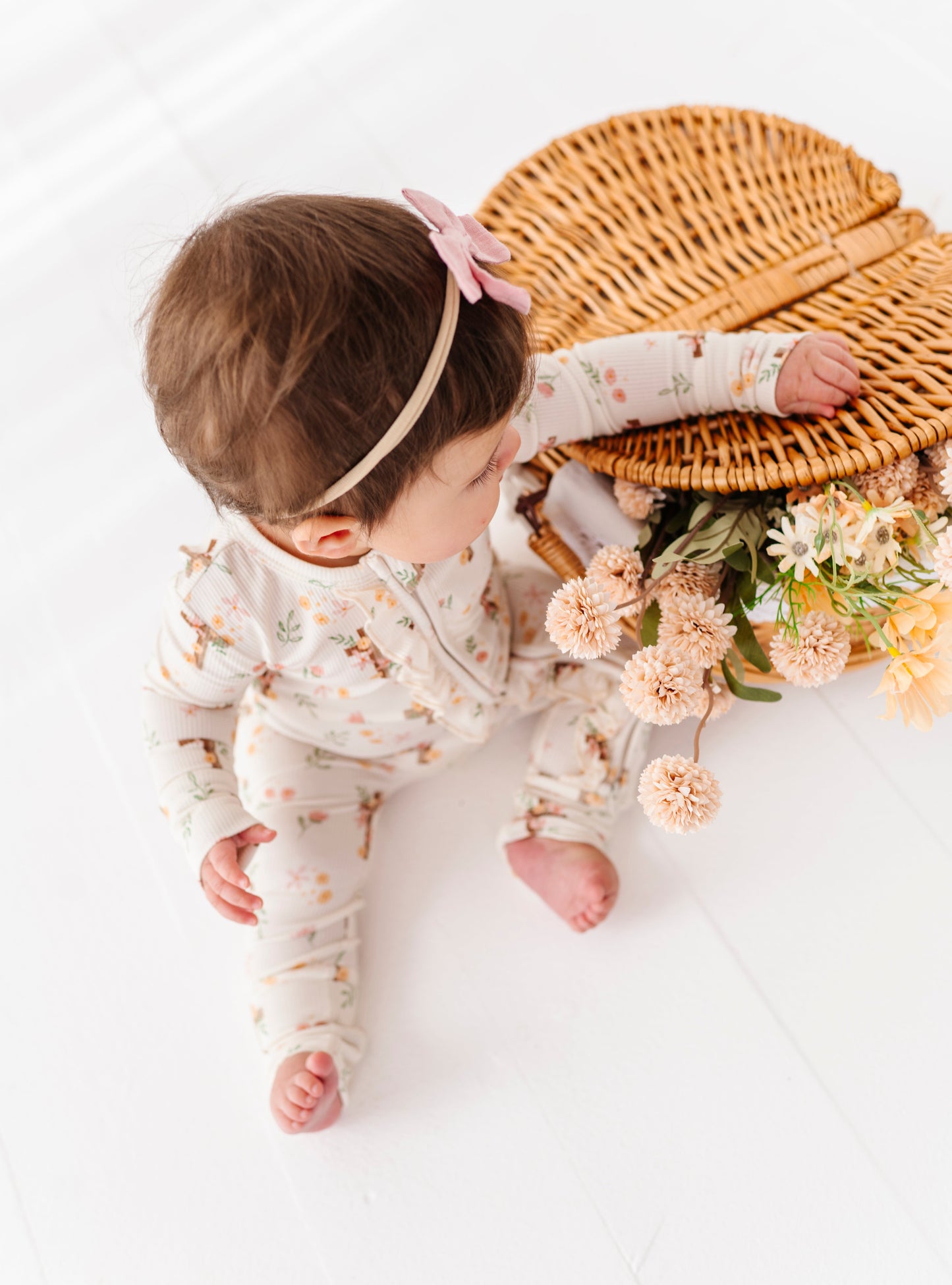 Baby in a floral bamboo convertible romper with a woven basket and flowers on a white background