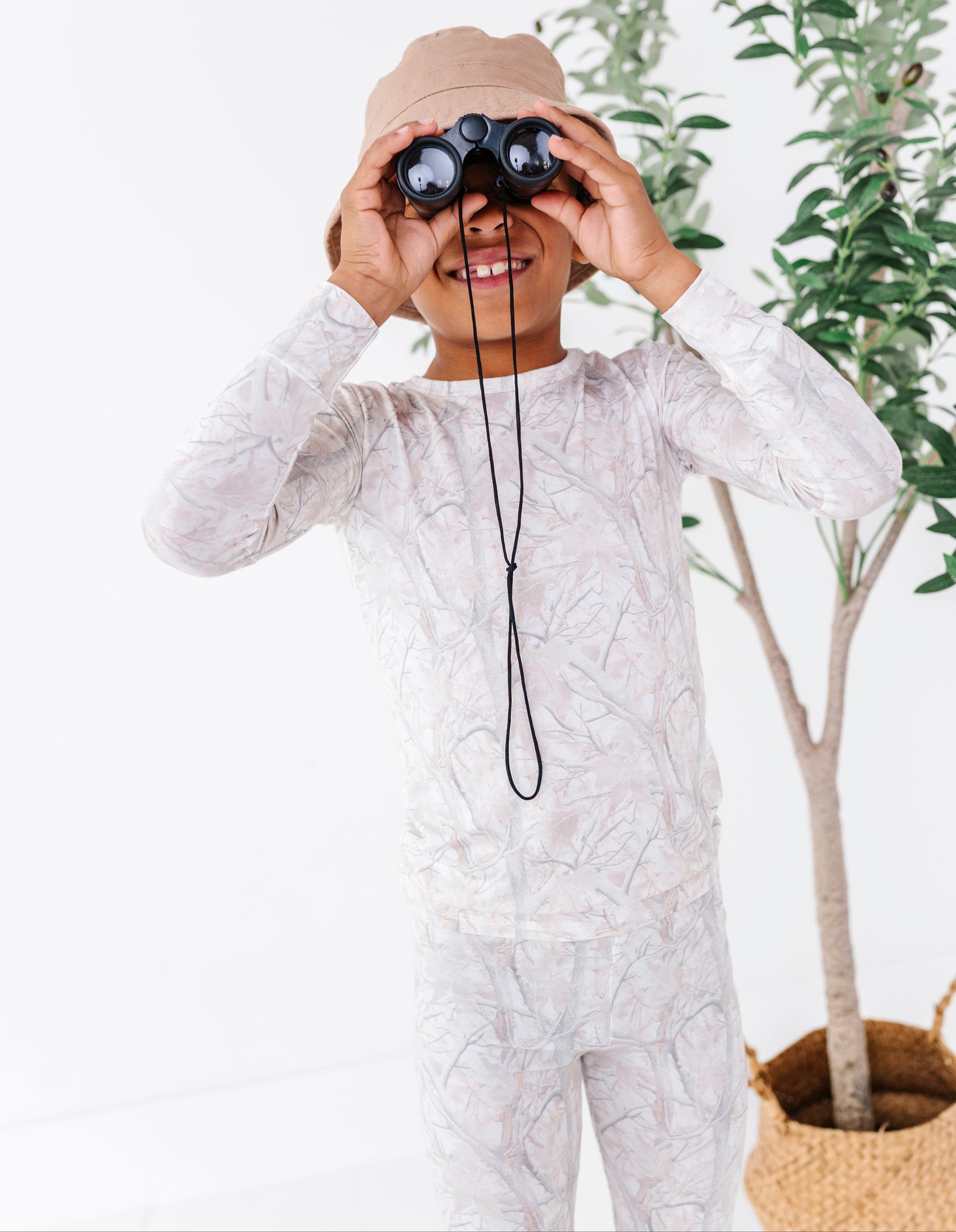 Child in a tree camo patterned outfit with binoculars, standing in front of a plant.