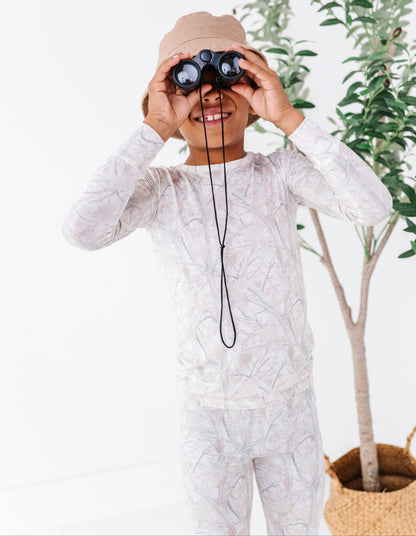 Child in a tree camo patterned outfit with binoculars, standing in front of a plant.