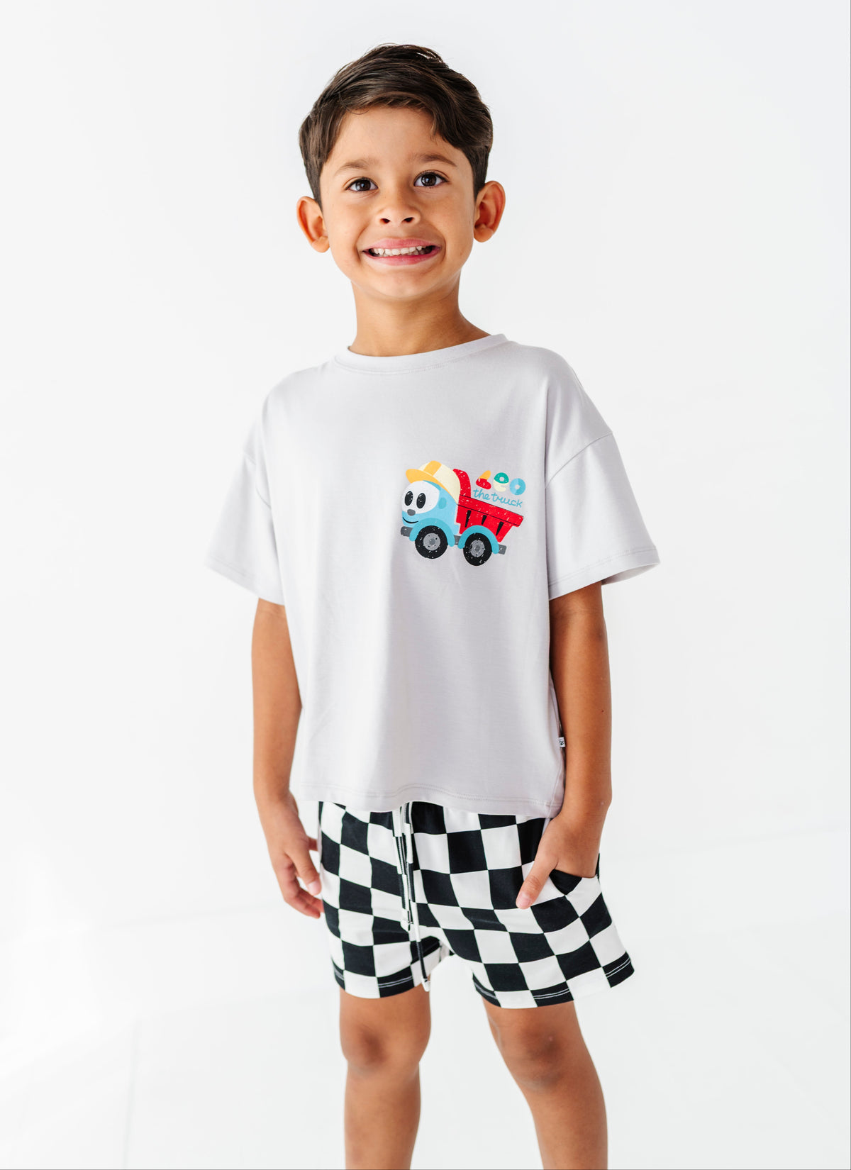 A young boy smiling, wearing a white short-sleeve t-shirt with a colorful dump truck graphic on the chest, paired with black and white checkered shorts, against a plain white studio background.