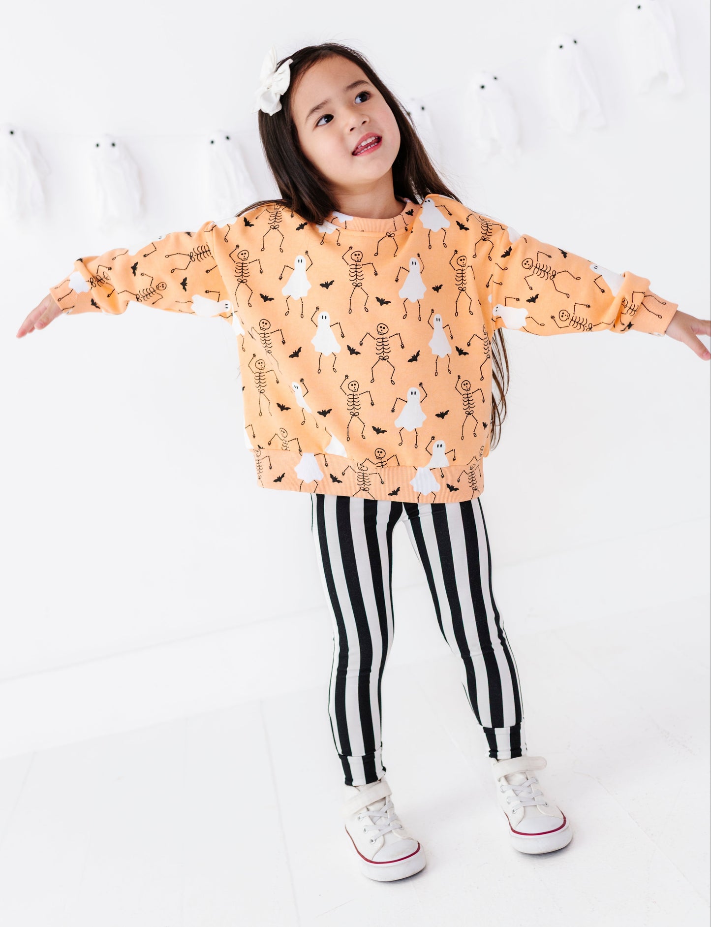 A young girl modeling a light orange long-sleeve hoodie with a black and white skeleton and ghost pattern, paired with black and white vertically striped leggings and white sneakers, posing with outstretched arms against a white wall decorated with small white ghosts.