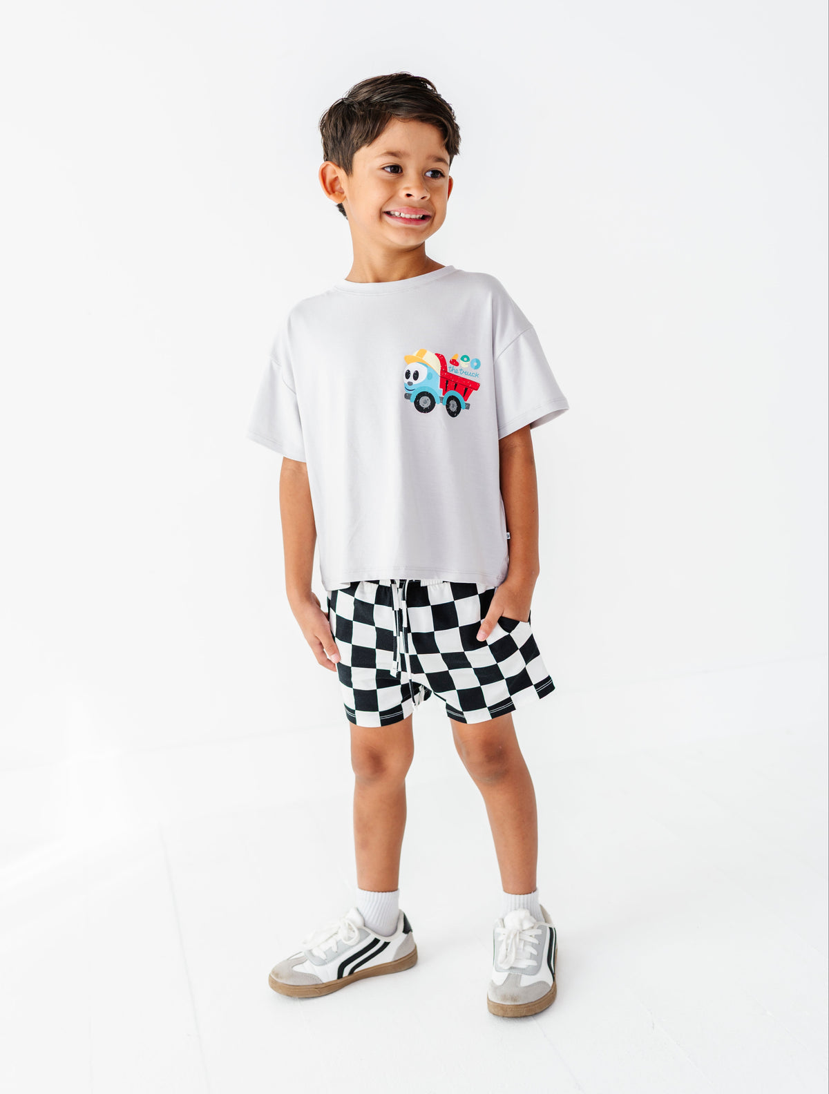 A young boy stands against a plain white studio background wearing a white short-sleeve t-shirt with a small colorful truck graphic on the chest, black-and-white checkered shorts, white socks, and white sneakers.