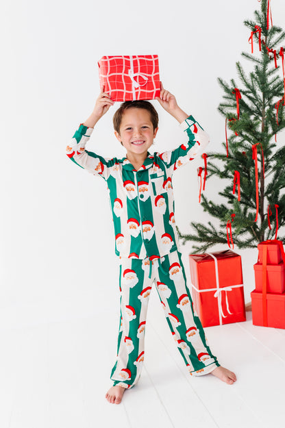 A young boy wearing a green and white Santa Claus patterned bamboo pajama set stands barefoot on a white floor, holding a red wrapped gift box above his head. A decorated Christmas tree and red gift boxes are visible in the background.