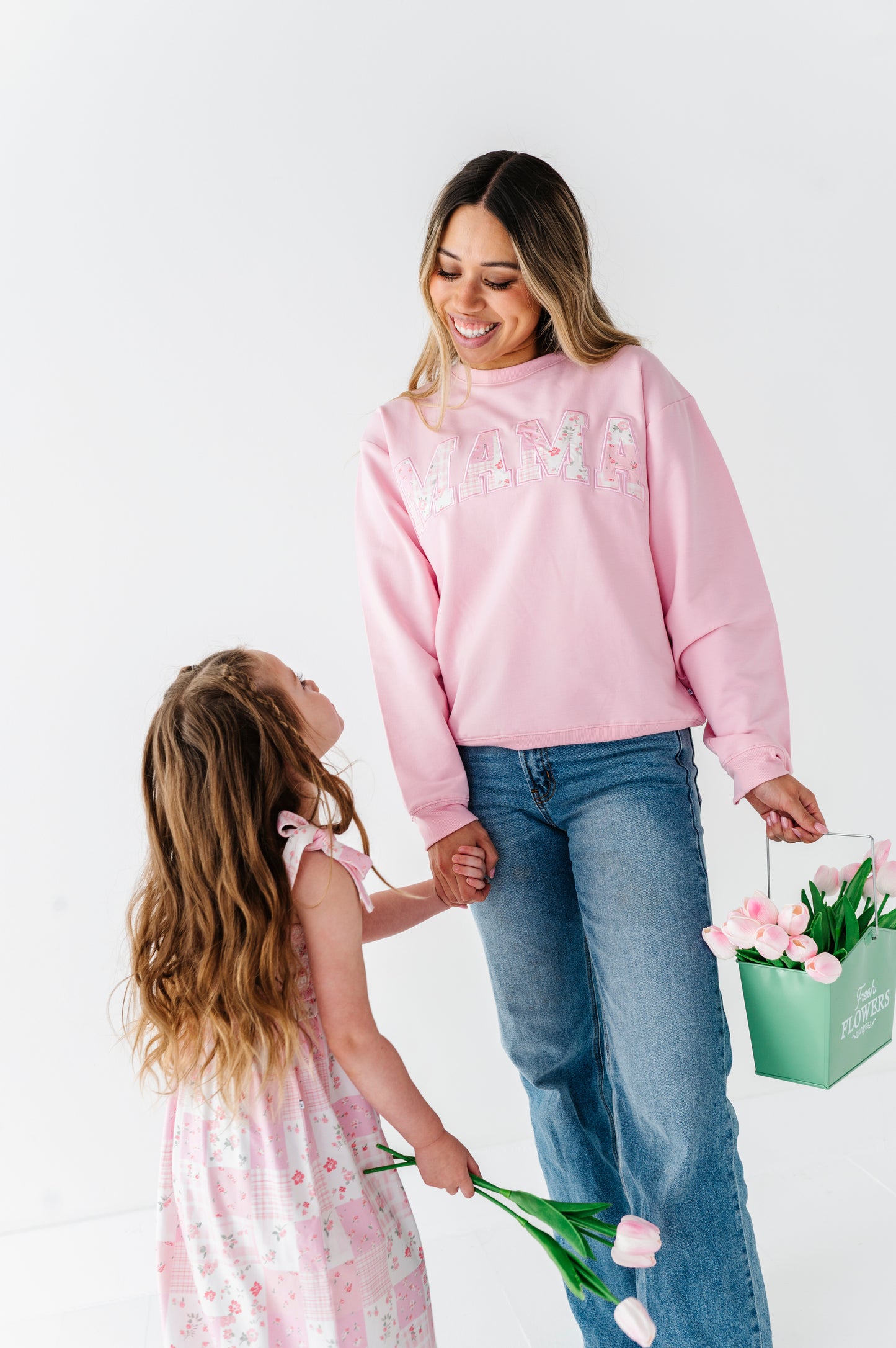Woman in pink bamboo-cotton sweatshirt and jeans holding hands with a young girl in a floral dress, both holding flowers against a white background.