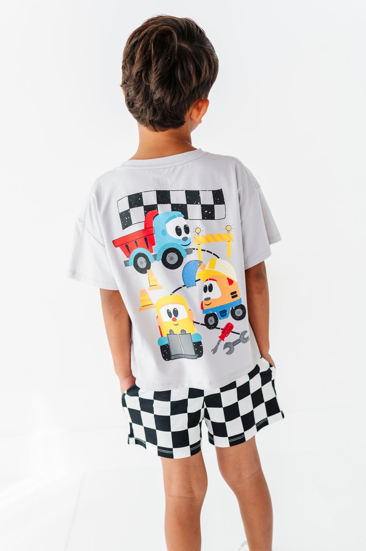 A child shown from the back wearing a white short-sleeve t-shirt with a colorful cartoon race truck graphic on the back and black-and-white checkered shorts, against a plain white studio background.