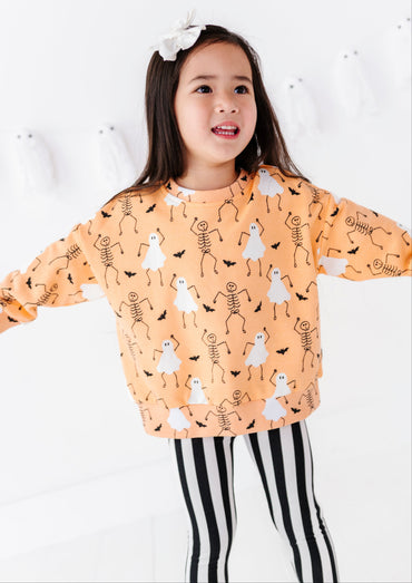 A young girl with a white bow in her hair is wearing a light orange long-sleeve sweatshirt with a black and white skeleton and ghost pattern, paired with black and white striped leggings, set against a white wall with ghost decorations.