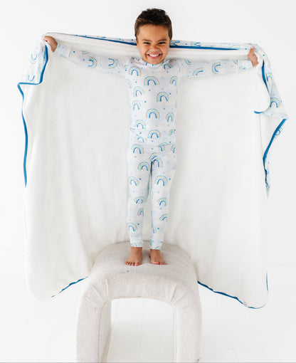 Child in bamboo pajamas with watercolor rainbow pattern holding a matching bamboo blanket with blue trim on a white background