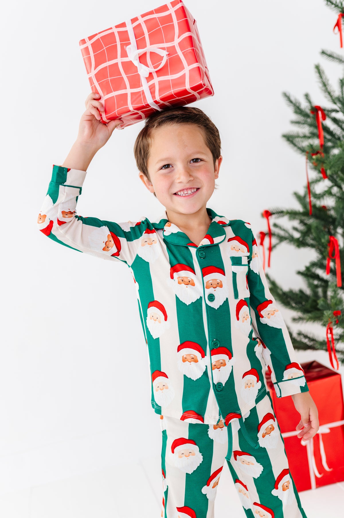 A young boy wearing festive green and white Santa Claus bamboo pajamas smiles while holding a red gift box balanced on his head, with a decorated Christmas tree and wrapped presents in the background.