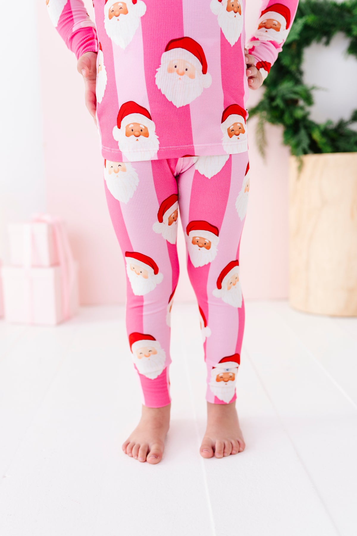 A child wearing a pink Santa Claus patterned bamboo pajama set, featuring a long-sleeve top and matching leggings, standing barefoot in a brightly lit room.