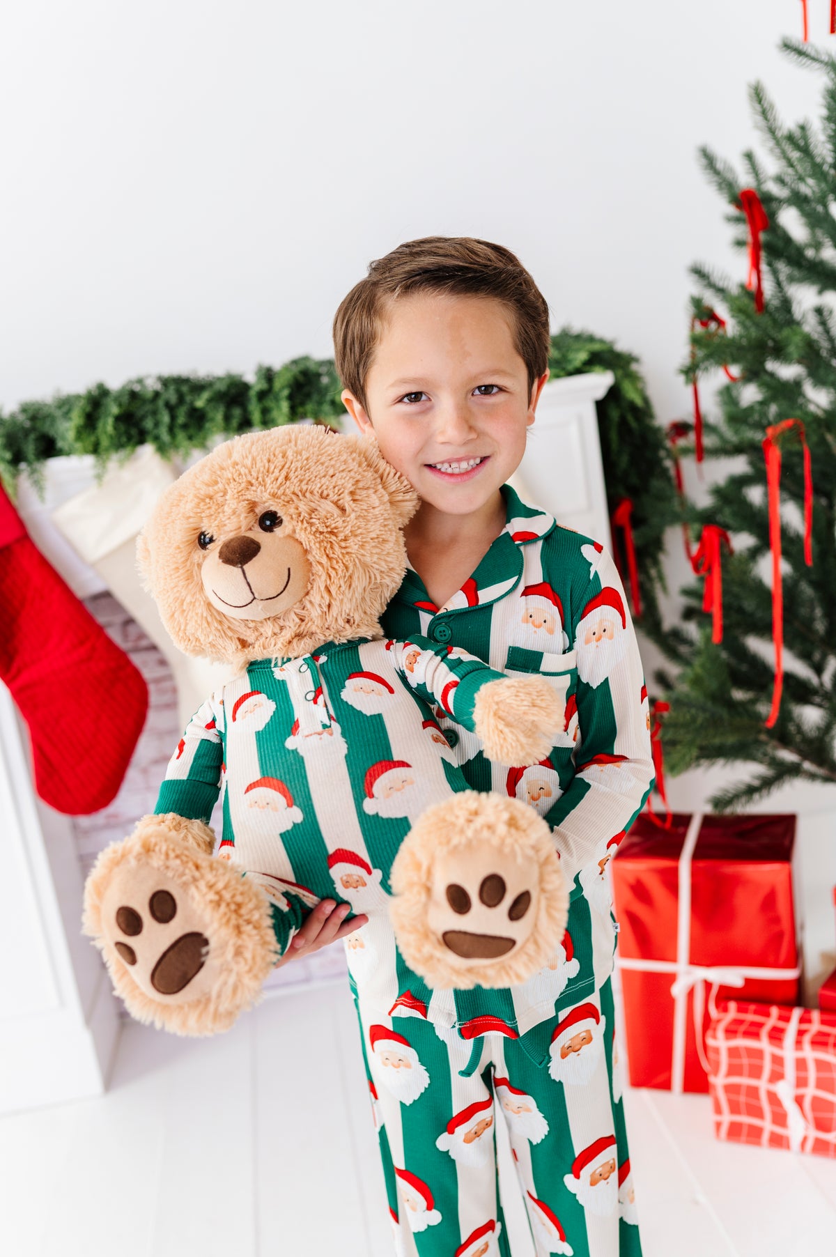 A young boy smiling while wearing festive green and white Santa bamboo pajamas, holding a large beige teddy bear and matching plush paw slippers in a decorated holiday room with a Christmas tree and wrapped presents.