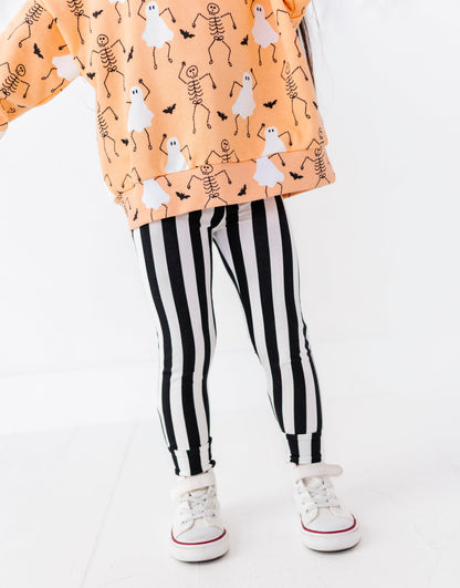 A child's lower body wearing black and white vertically striped leggings, a peach-orange sweatshirt with a white ghost and black skeleton print, and white sneakers with red stripes, shown against a plain white studio background.