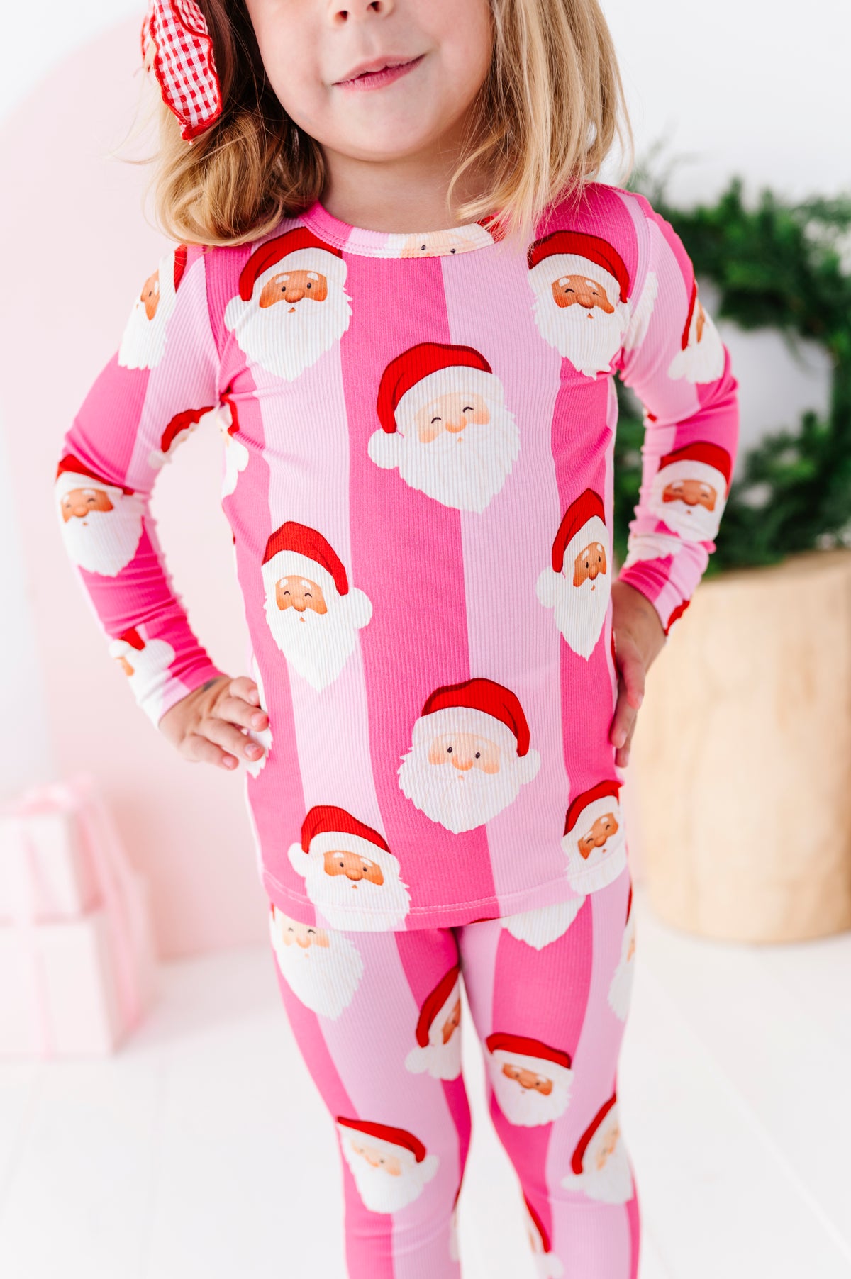 A young girl wearing a pink ribbed long-sleeve bamboo pajama set with a repeating Santa Claus face print, standing in a brightly lit indoor setting.