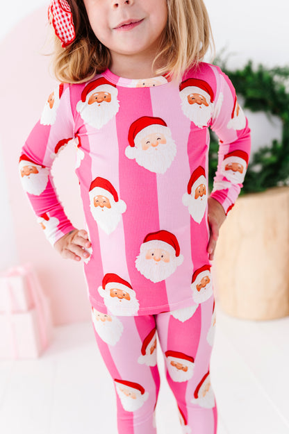 A young girl wearing a pink ribbed long-sleeve bamboo pajama set with a repeating Santa Claus face print, standing in a brightly lit indoor setting.