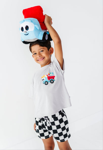 A young boy wearing a white t-shirt with a cartoon dump truck graphic and black-and-white checkered shorts, balancing a plush dump truck on his head against a white studio background.