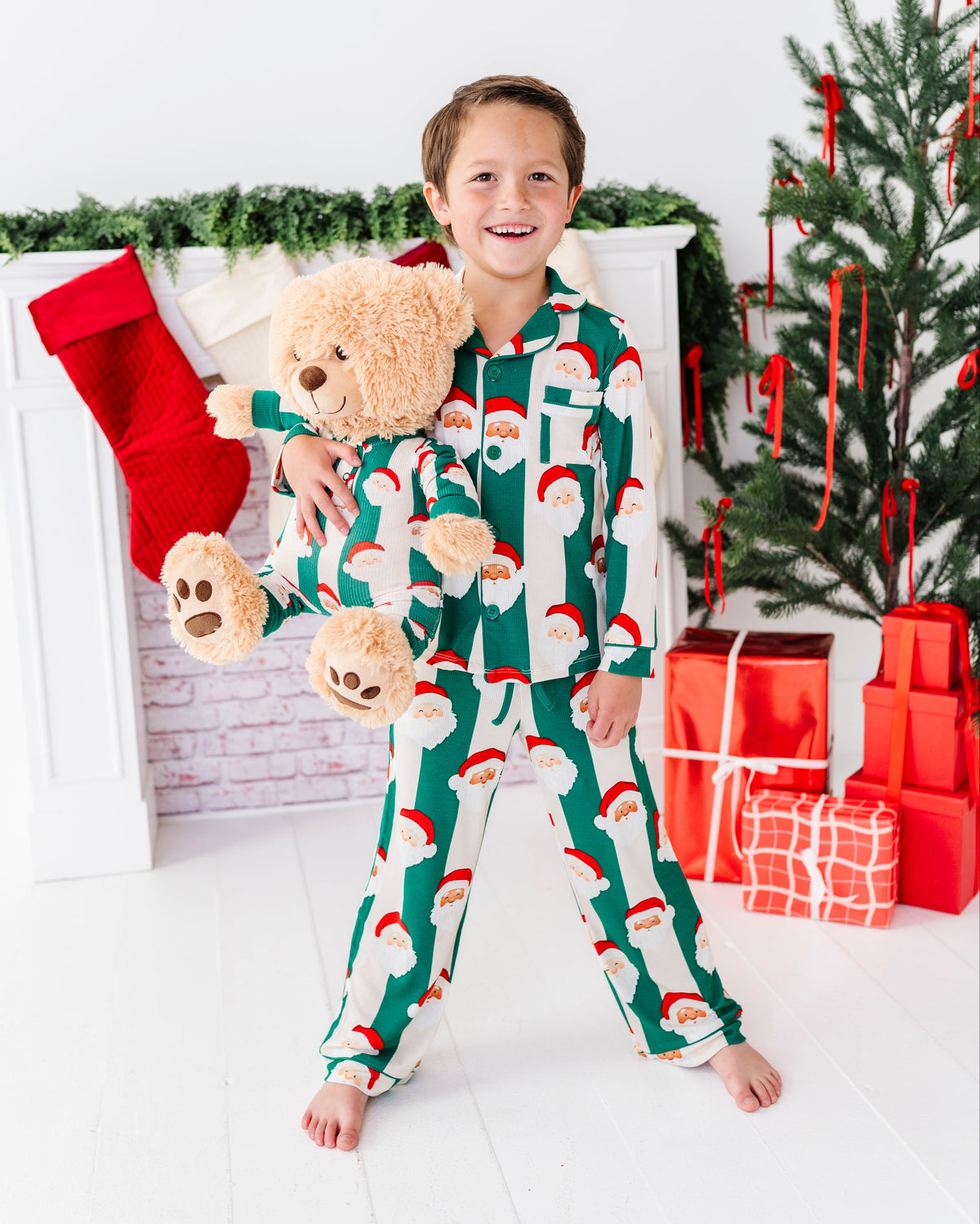 A young boy stands barefoot in a holiday-themed room, wearing a green and white Santa Claus patterned pajama set and holding a beige teddy bear. A Christmas tree, stockings, and wrapped gifts are visible in the background.